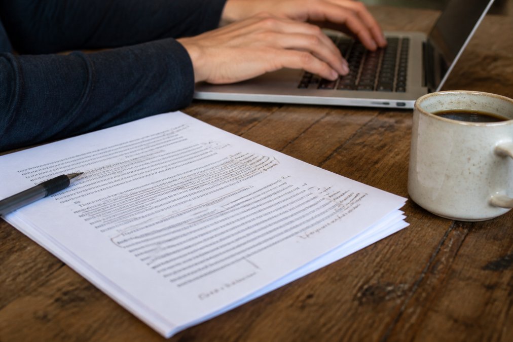 Close-up of a writer's hands typing on a laptop beside a printed manuscript with handwritten margin notes and a coffee cup on a wooden desk