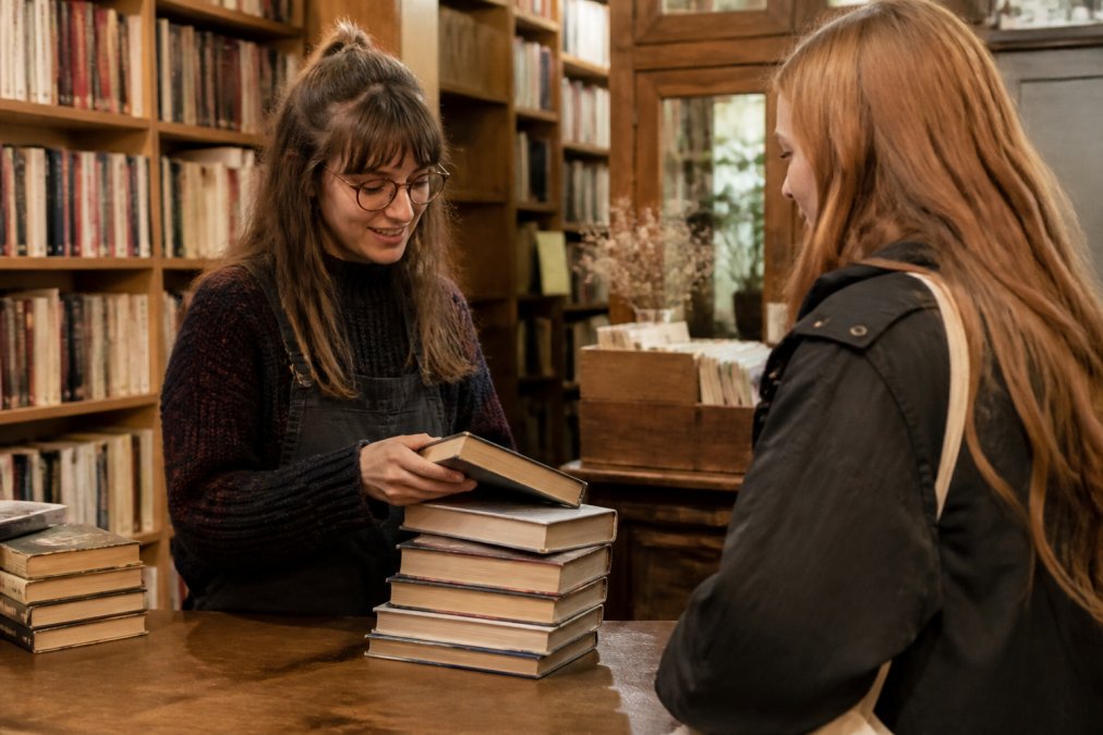 Friendly bookstore employee appraising a stack of books for a customer looking to sell used books in a cozy shop