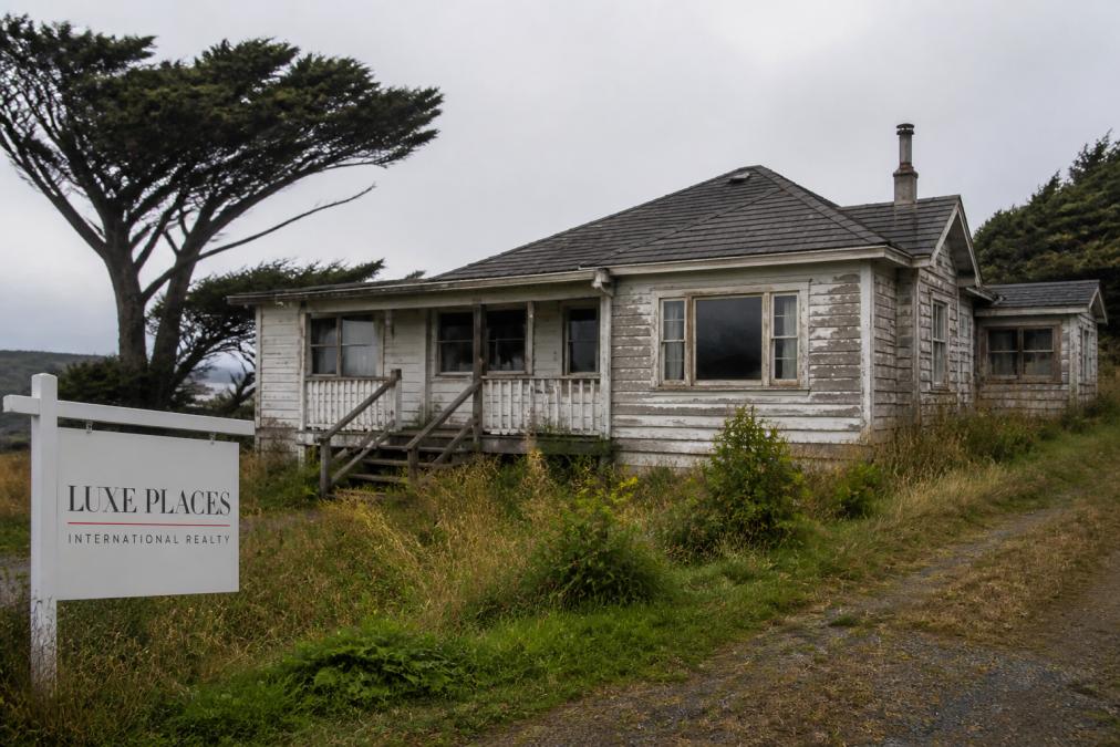 A weathered single-family home with peeling paint and an overgrown yard in a rural Northern California setting, with oak trees in the background and overcast coastal light