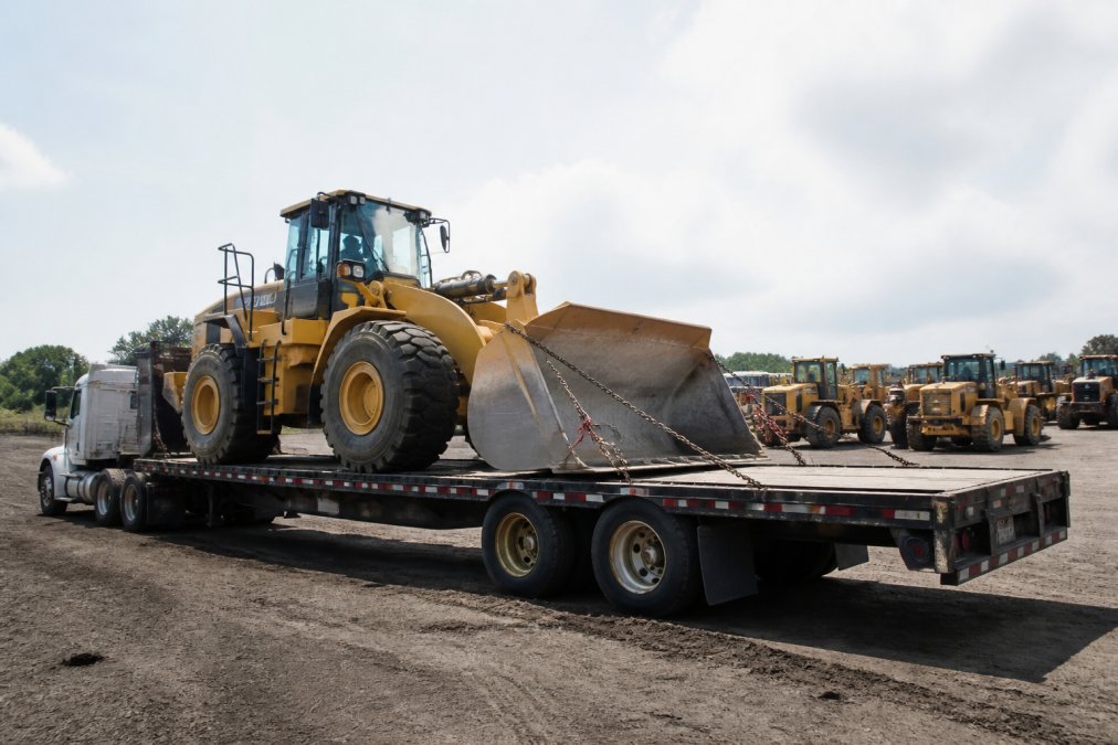 Industrial flatbed truck loaded with heavy machinery secured with chains, positioned at an equipment staging yard ready for transport