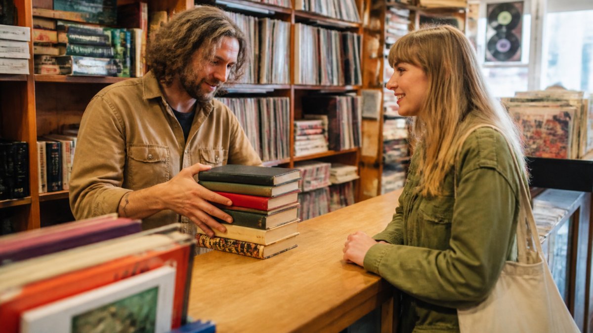 A friendly bookstore employee at a wooden counter examining a stack of used hardcover books while a customer smiles nearby, surrounded by shelves of vintage vinyl and paperbacks in a warm, nostalgic shop