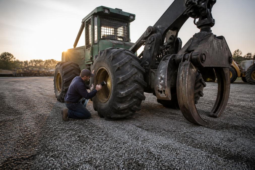 A used grapple skidder parked in a well-lit gravel yard at golden hour, a person in work clothes kneeling to inspect the undercarriage and tire tread while holding a flashlight