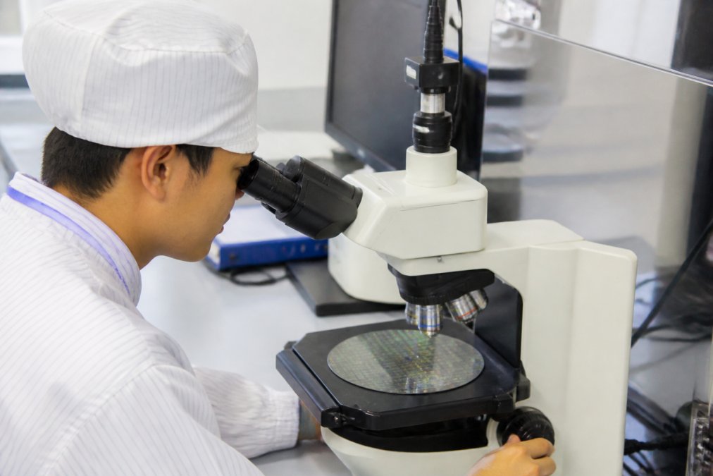 Technician reviewing wafer mapping data on high-resolution monitor in semiconductor fab control room, with multiple data screens displaying process parameters and color-coded defect distributions