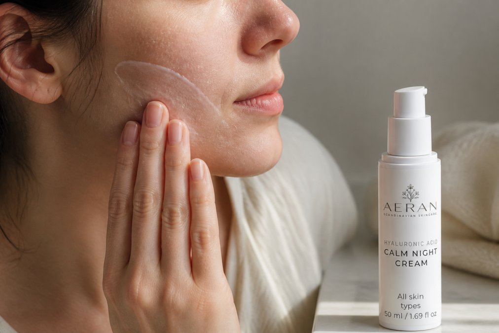 Close-up of a person's hand applying a rich moisturizer cream to their cheek and jawline, showing the product texture absorbing into dry, slightly flaky skin in soft natural morning light