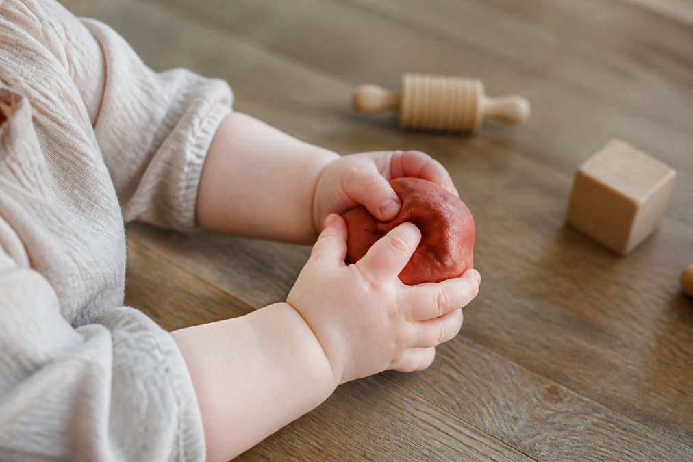 Babyhandjes knijpen voorzichtig in een bal rode natuurlijke klei op een houten tafel met speelgoed op de achtergrond