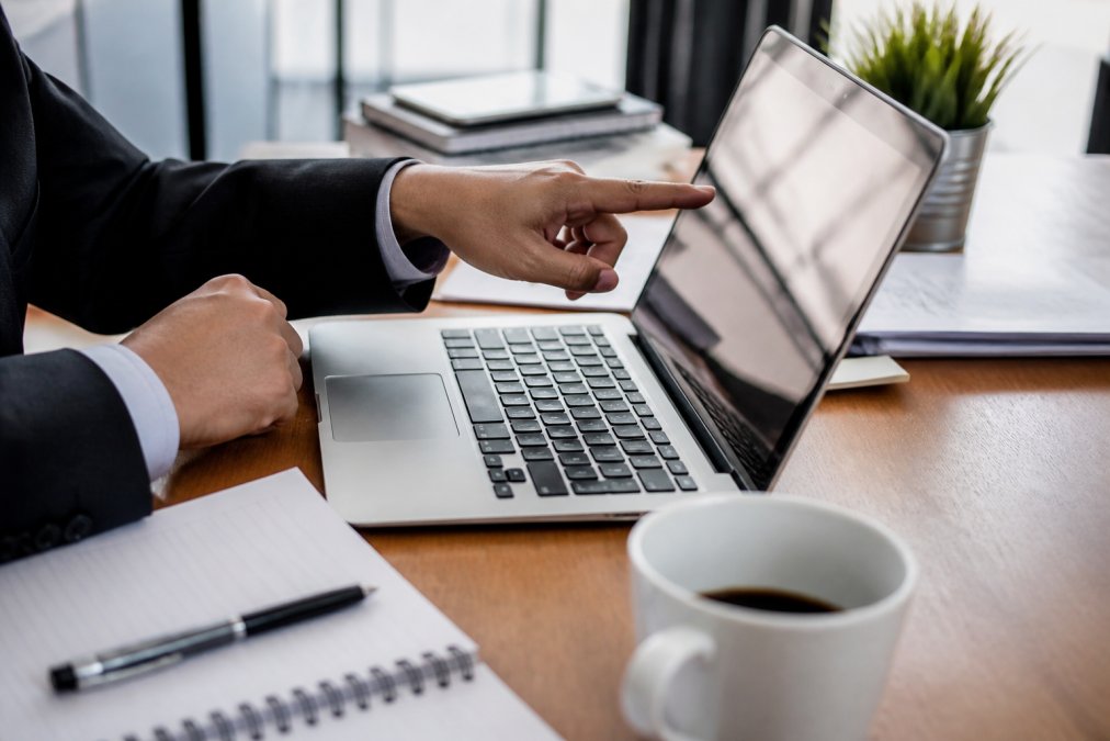 Close-up of a business professional's hands pointing at performance metrics on a laptop screen, with a notepad and coffee cup on the desk beside them