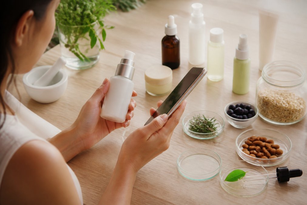Close-up of hands holding a skincare bottle while reviewing product information on a smartphone, with other beauty products and ingredients visible nearby on a light wooden surface