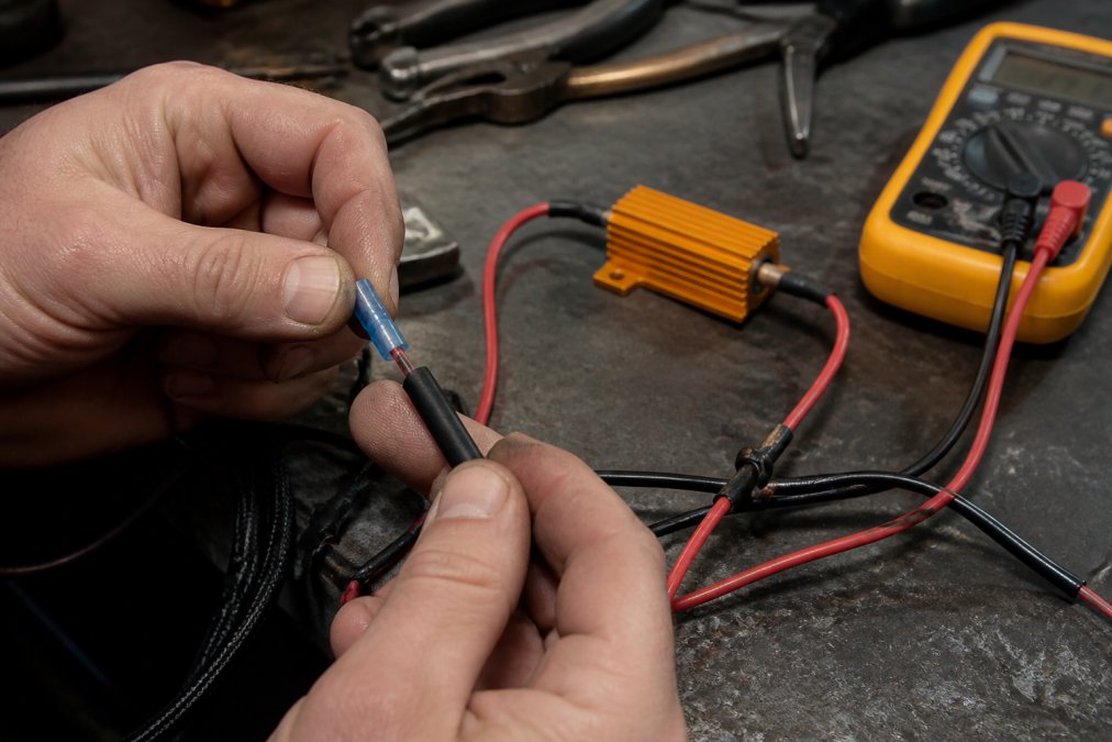 Close-up of hands using a crimp connector and heat shrink on a load resistor wired into a car's indicator circuit, with a multimeter resting nearby on a garage workbench
