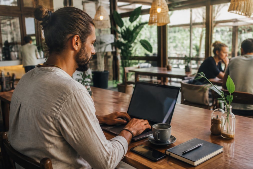 Digitaler Nomade arbeitet an seinem Laptop in einem modernen Café und verwaltet mehrere Online-Geschäftsbereiche auf seinem Monitor