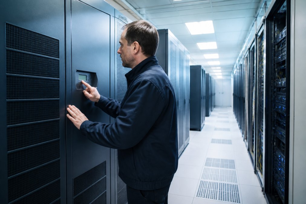 A technician in a data centre inspecting the control panel of a modern precision cooling unit, with rows of server racks visible in the background under cool blue lighting