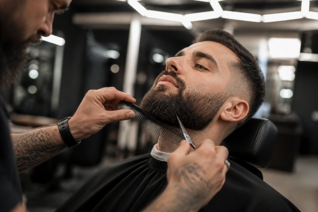 Un barbier haut de gamme sculpte une barbe au peigne et aux ciseaux dans un salon moderne et lumineux.