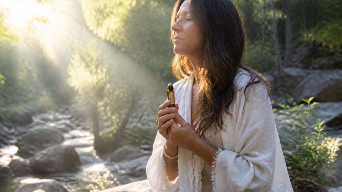 Person sitting peacefully in a serene natural setting at sunrise, holding a crystal essential oil roller bottle in their hands, with soft golden light streaming through trees and a calm, grounded expression on their face