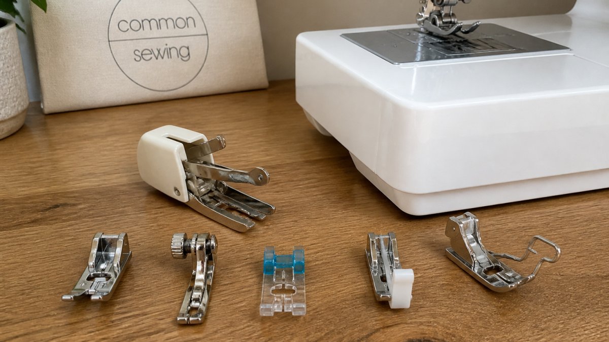 Close-up overhead shot of 6-8 different presser feet arranged on a wooden surface next to a sewing machine, showing the distinct shapes and designs of standard, walking, zipper, blind hem, and gathering feet