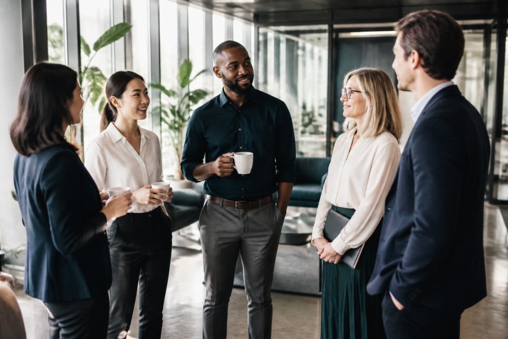 A diverse group of professionals chatting in a bright modern coworking space with floor-to-ceiling windows and plants, some holding coffee cups, engaged in conversation