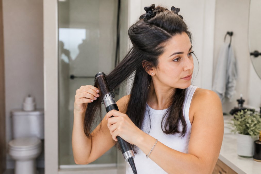 Woman with thick, voluminous hair using an air styler on a lower section of hair, with clips holding up the remaining sections, demonstrating proper sectioning technique in natural bathroom lighting