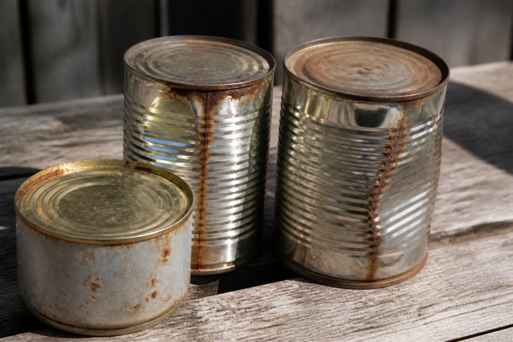 Close-up of damaged metal cans showing rust spots, dents along the seams, and slight bulging on the sides, arranged on a weathered wooden surface in natural afternoon light