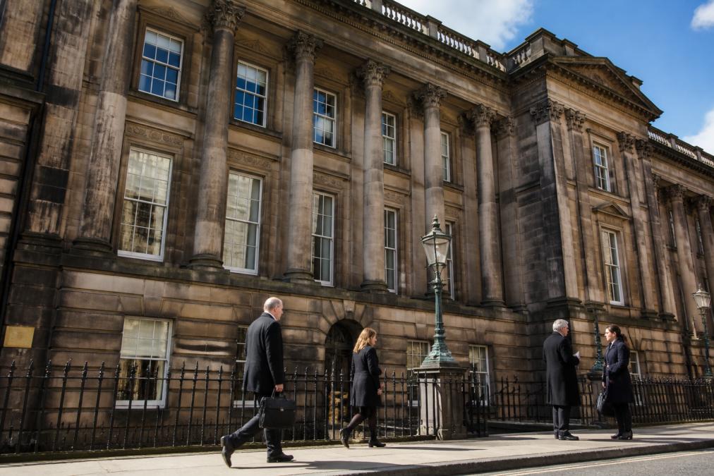 A wide exterior photograph of a historic Scottish court building with classical stone architecture, people walking past on a pavement, and a clear sky overhead