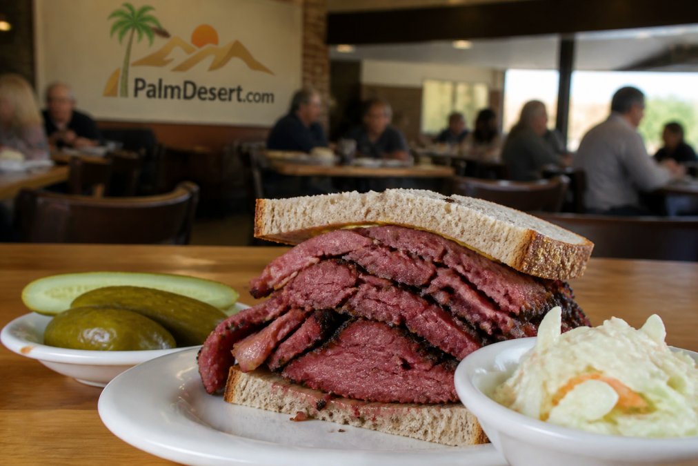 A close-up of a towering pastrami sandwich on rye bread with a side of pickles and coleslaw on a deli table, warm lighting, diners blurred in the background