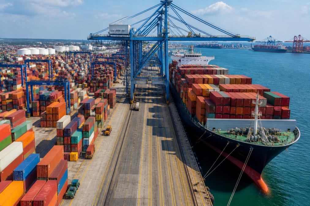 Busy container port terminal with stacked shipping containers, cargo ships docked at berths, and automated port equipment moving containers under bright daylight
