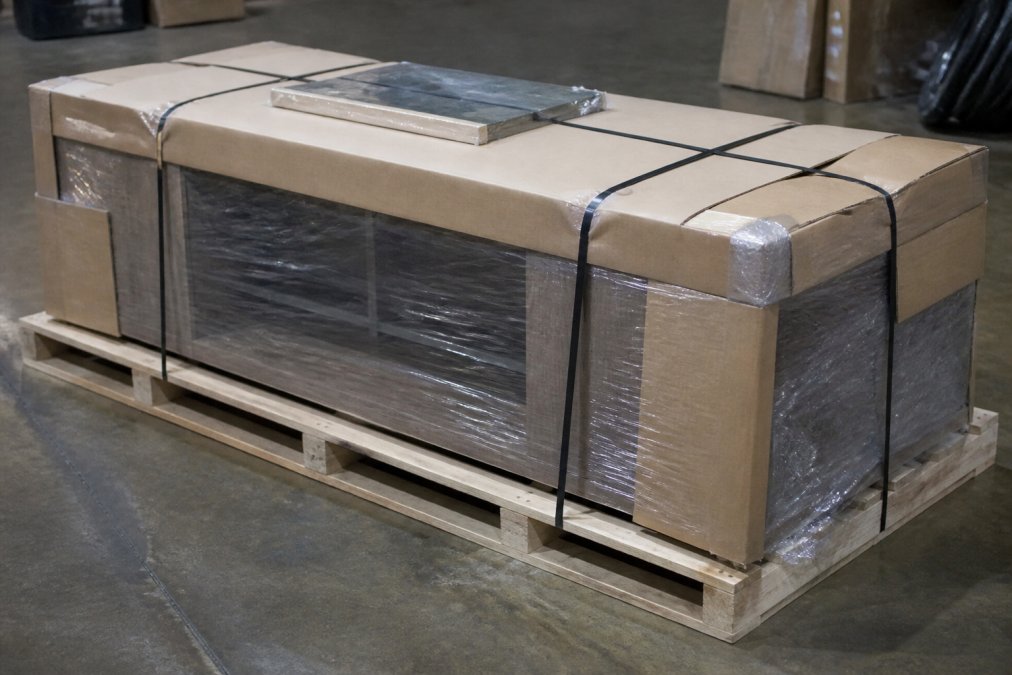 Close-up of a wooden pallet with a wrapped outdoor cooking table secured with heavy-duty straps, showing proper crating and protective cardboard covering for freight shipment