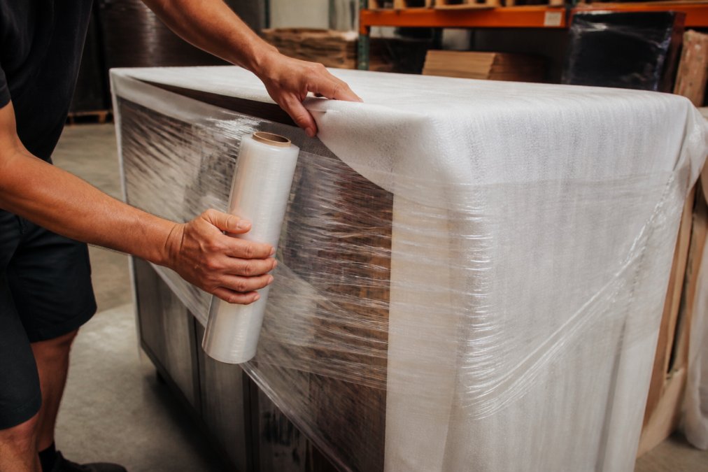 Close-up of hands carefully wrapping a heavy furniture piece with protective foam padding and securing it with clear plastic stretch film in a well-lit workspace