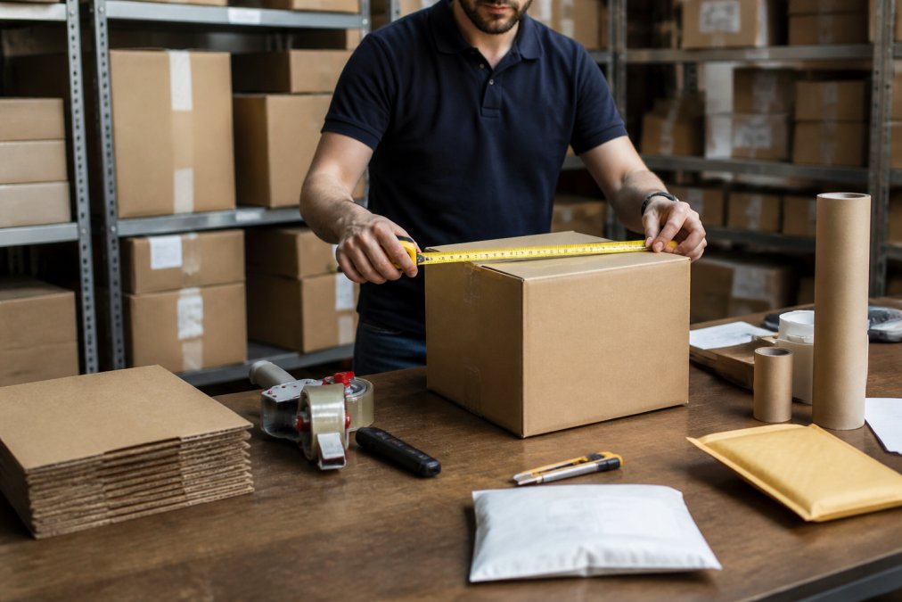 Logistics professional measuring a cardboard box with a tape measure at a warehouse workstation, with shipping labels and packaging materials visible on the desk