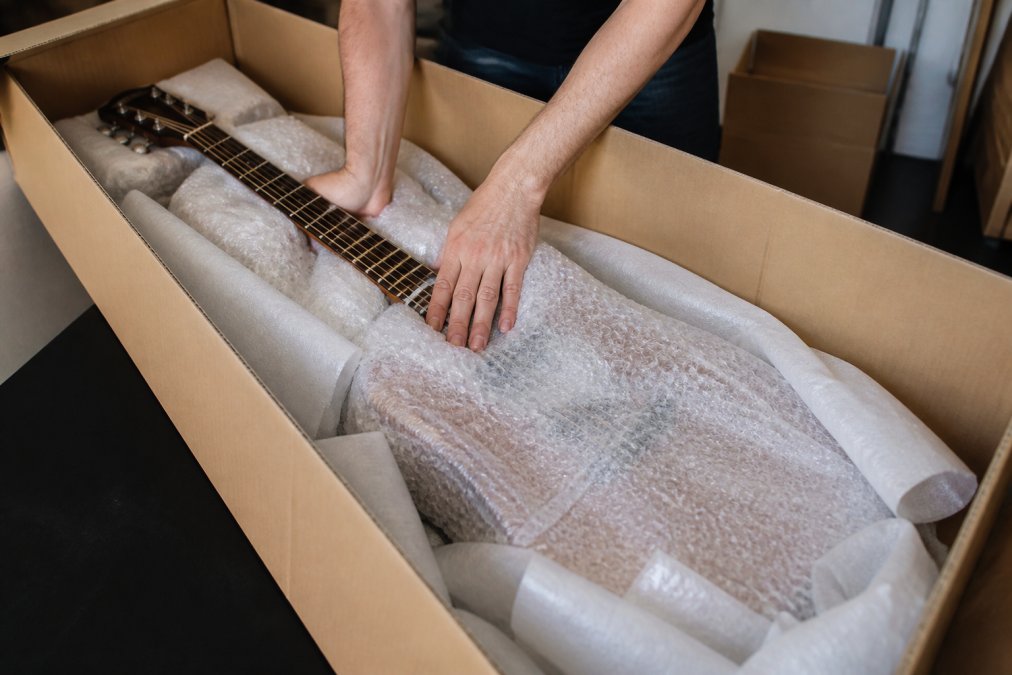 Close-up of someone wrapping a guitar with bubble wrap and protective padding in a cardboard box, showing proper layering technique with the instrument positioned diagonally for stability