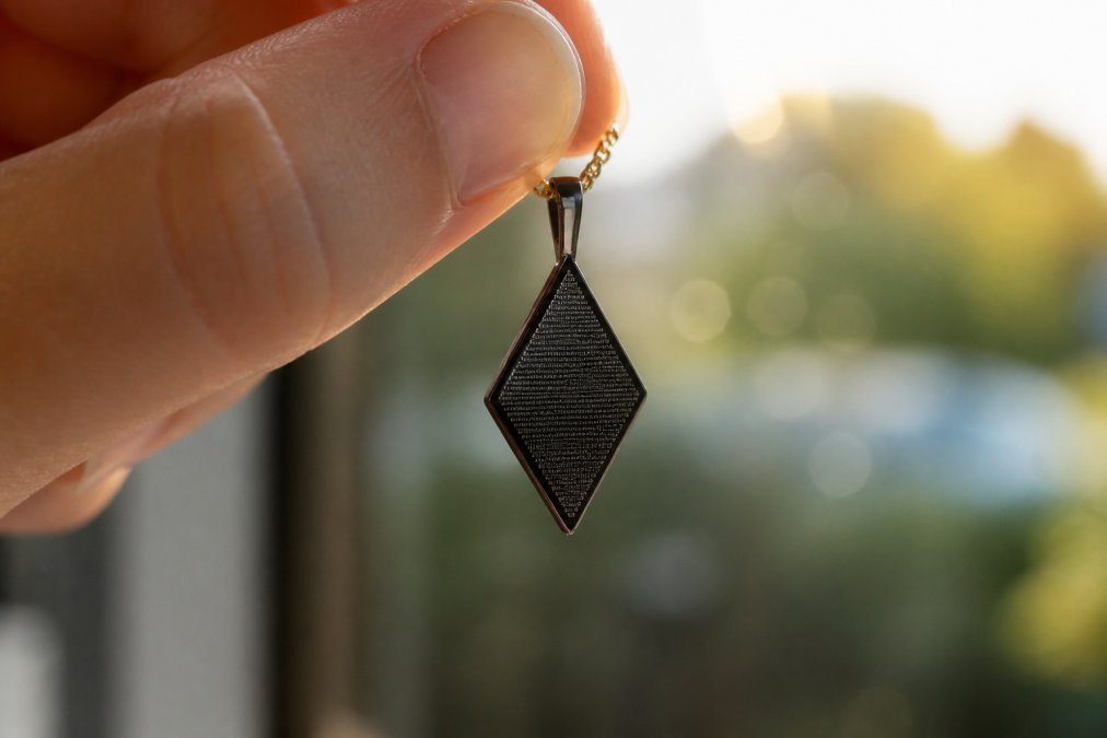 Close-up of a nano-engraved Bible chip pendant being held up to light, showing the precision of microscopic engraving on the black diamond-shaped charm with a delicate gold chain, detailed surface texture visible