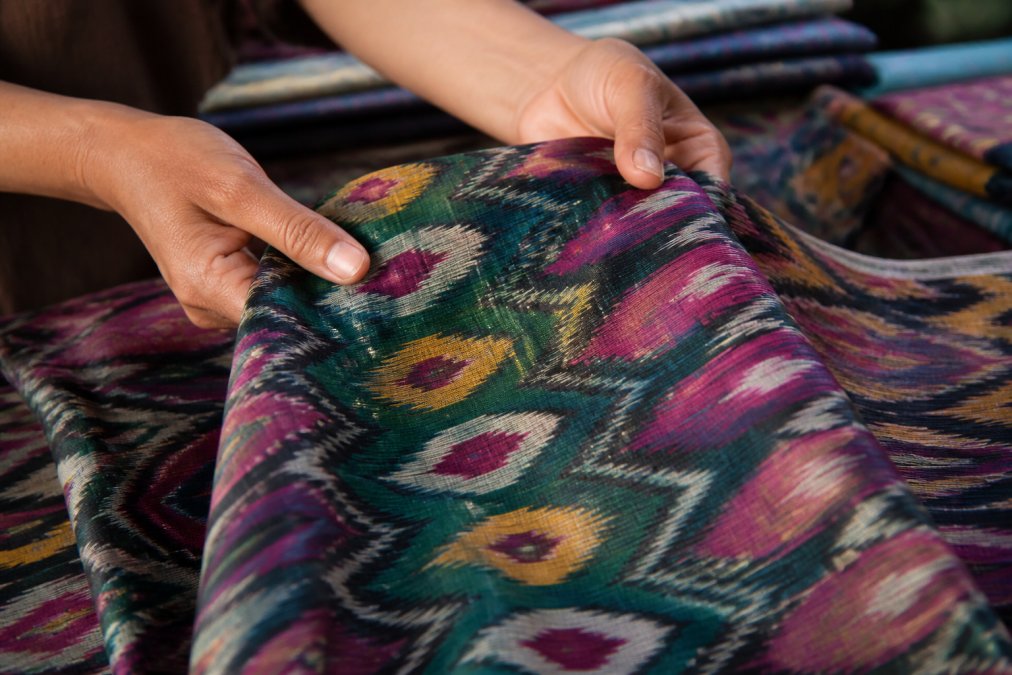 Close-up of hands holding woven ikat fabric with intricate geometric patterns in jewel tones, showing the blurred edges and layered color variations characteristic of traditional resist-dyeing techniques