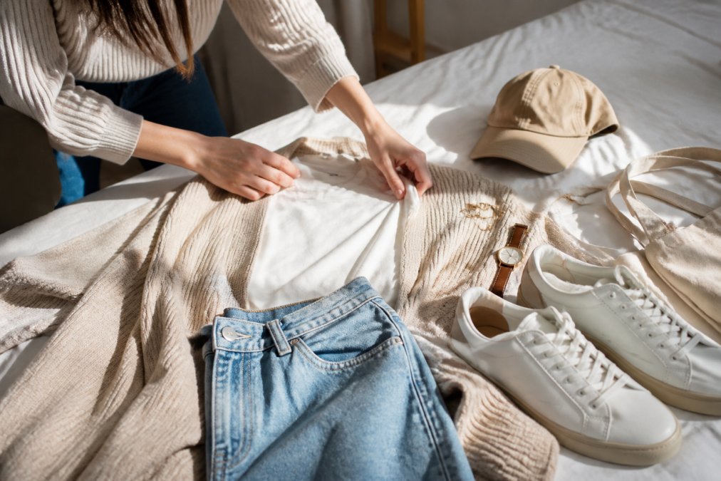 Close-up of hands styling an outfit on a bed, showing layering techniques with neutral basics, sneakers, and accessories arranged together in natural light