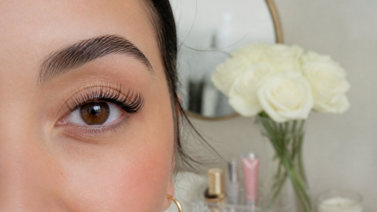 Close-up of a woman's eye wearing featherlight false lashes with a natural, wispy finish, soft daylight, minimal makeup, elegant vanity setting in the background