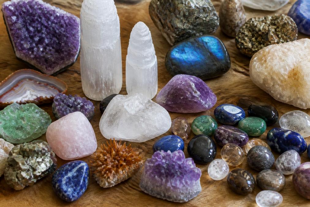 Close-up display of various raw and polished crystals, gemstones, and minerals arranged on a wooden surface, showing different colors, shapes, and textures including amethyst, selenite, and other specimens under natural lighting