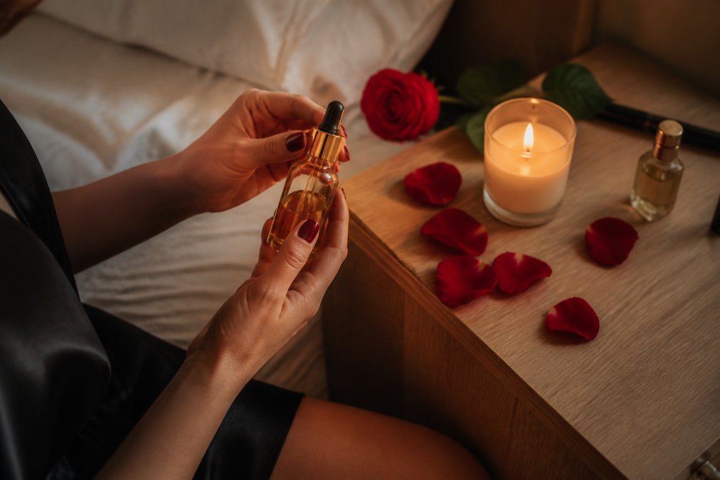 A close-up of a woman's hands holding a small elegant bottle of intimate oil beside rose petals and a lit candle on a wooden nightstand, warm and romantic lighting