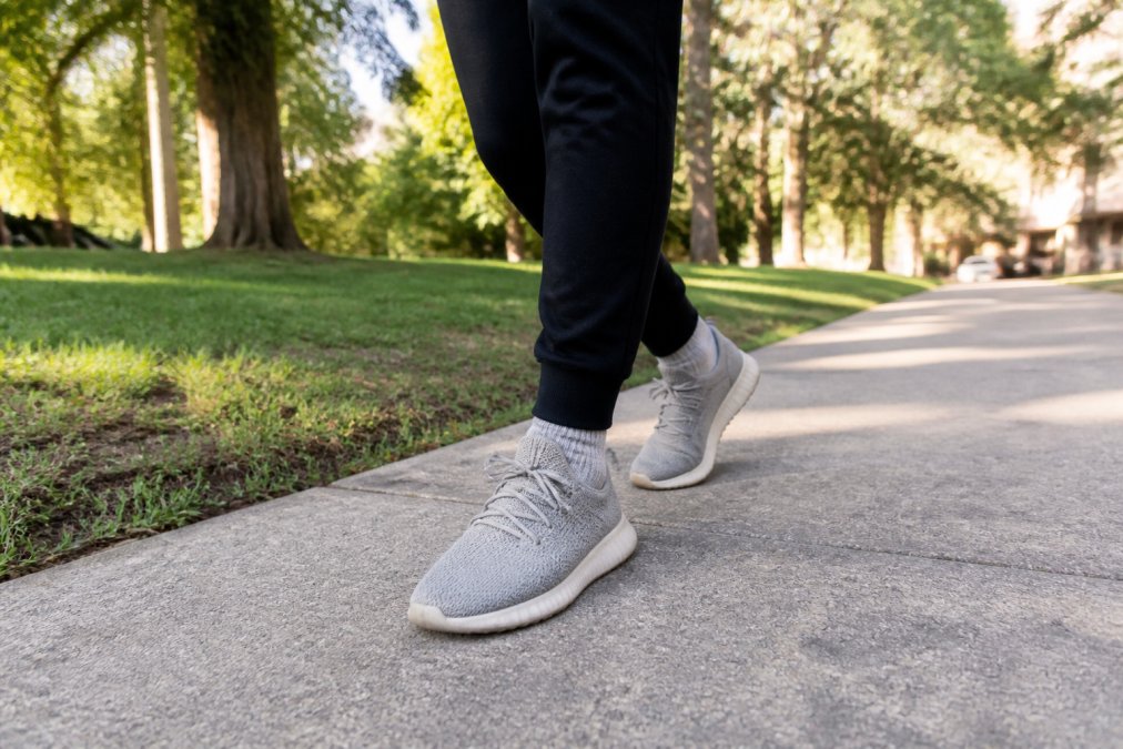 Person walking outdoors on tree-lined path wearing casual sneakers, natural daylight showing shoe in everyday use on concrete and grass terrain