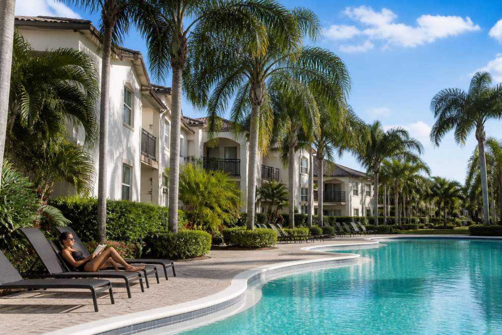 A modern garden-style apartment building in Boca Raton with palm trees lining the walkway, a resident relaxing by the resort-style pool in the foreground, clear blue sky overhead