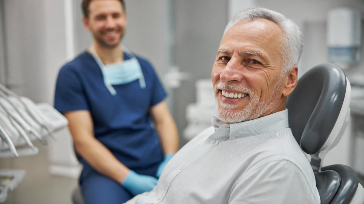 Older adult smiling confidently in a dental treatment chair with natural clinical lighting, dentist visible in the background
