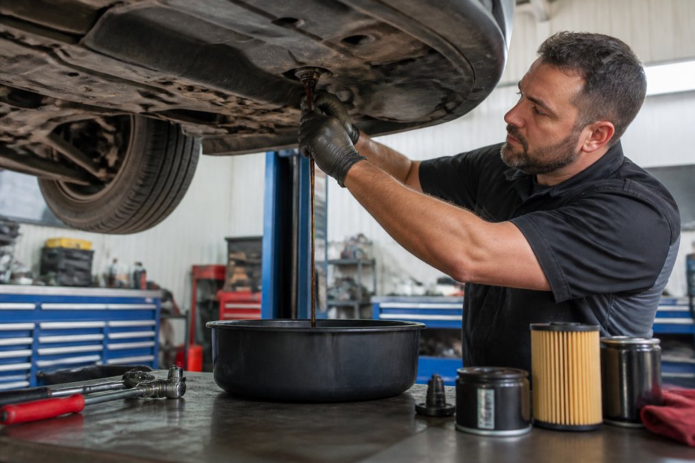 Professional mechanic performing oil change on a vehicle in a well-lit workshop, draining old oil into a container with diagnostic tools and spare filters visible on the workbench