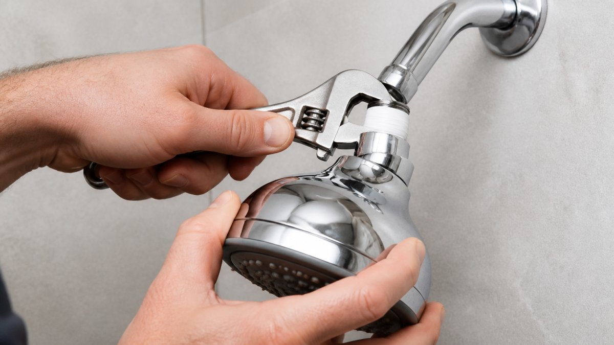 Professional plumber's hands holding an adjustable wrench while removing a chrome shower head from a shower arm, showing the connection point and thread seal tape wrapped around the threads