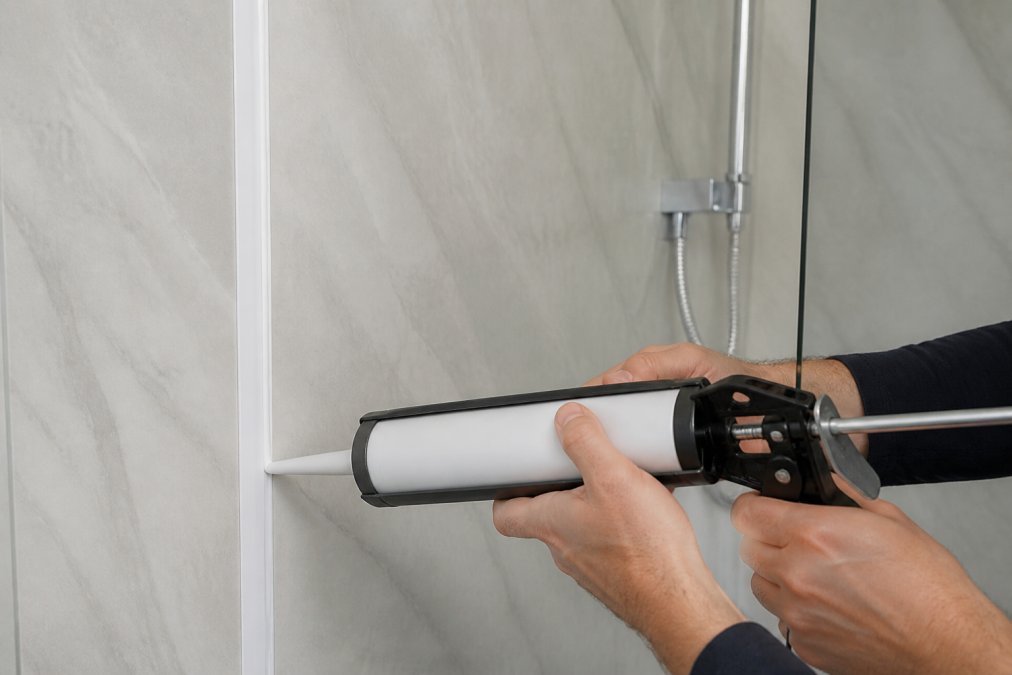 Hands applying white silicone sealant along a trim joint on shower wall panels using a caulking gun, showing proper sealing technique in a modern wet room with natural lighting