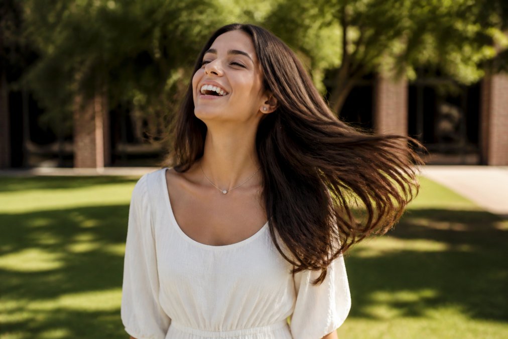 A woman with long, flowing brunette hair laughing and gently tossing her head, showing the natural swing of the hair in soft, warm natural light