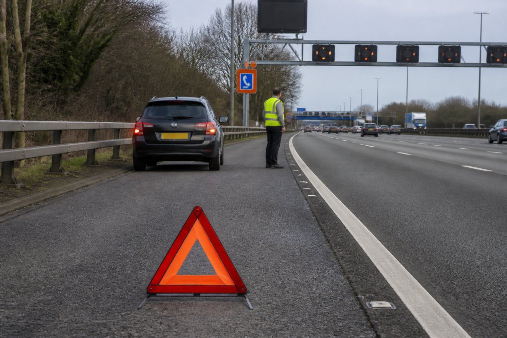 Signal for Help on Smart Motorways: A Driver’s Guide Stationary vehicle on smart motorway with hazard lights activated, emergency warning triangle positioned 45 metres behind it, driver in high-visibility vest standing safely on hard shoulder near emergency refuge area sign, with overhead gantry signals visible