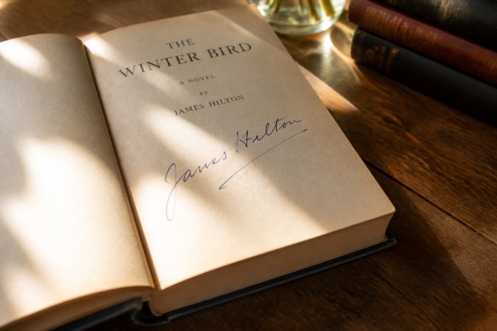 Close-up of an open signed vintage book with handwritten author signature visible on title page, placed on a wooden table with warm afternoon sunlight streaming across aged pages, suggesting heritage and personal connection