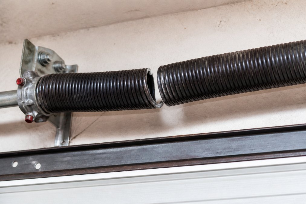 Close-up of a broken torsion spring mounted above a garage door, showing a clear gap between separated coils with a jagged metal fracture point, photographed in bright daylight