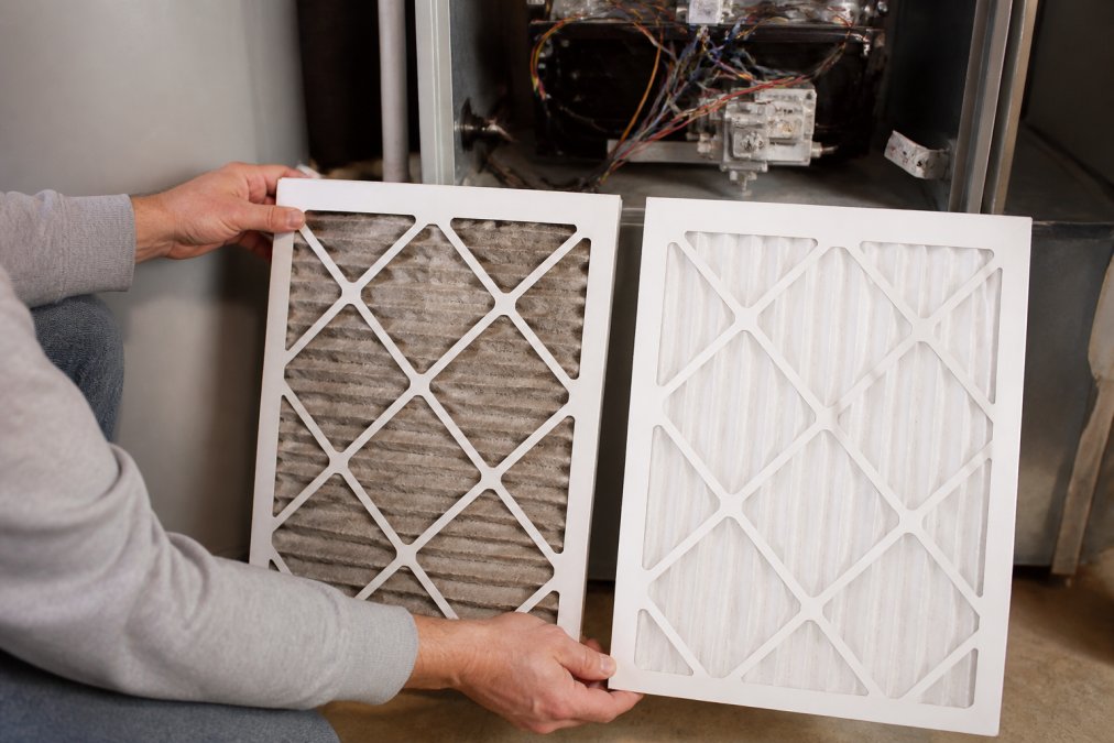 Homeowner's hands holding a visibly dirty air filter next to a clean replacement filter in front of an open HVAC unit compartment