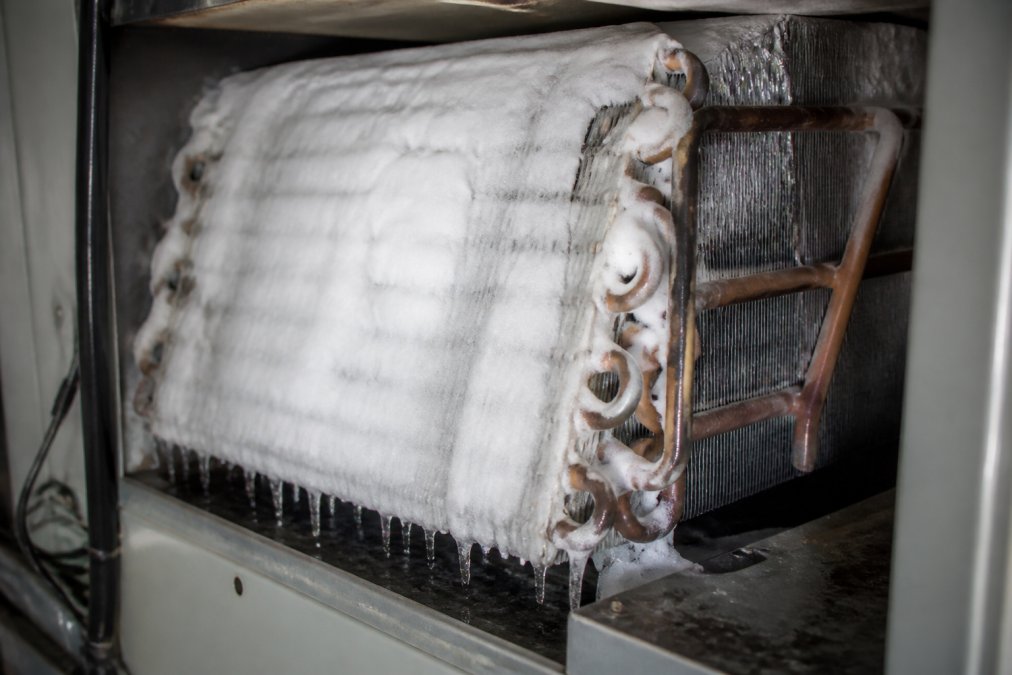 Close-up of a residential AC evaporator coil covered in thick white ice, with copper tubing visible and frost forming on the metal fins inside the air handler cabinet