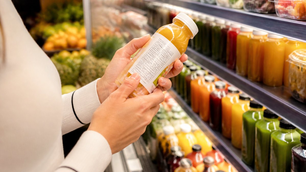 Close-up of a person's hands holding a juice bottle and turning it to read the ingredient list in a bright grocery aisle, with fresh fruit blurred in the background