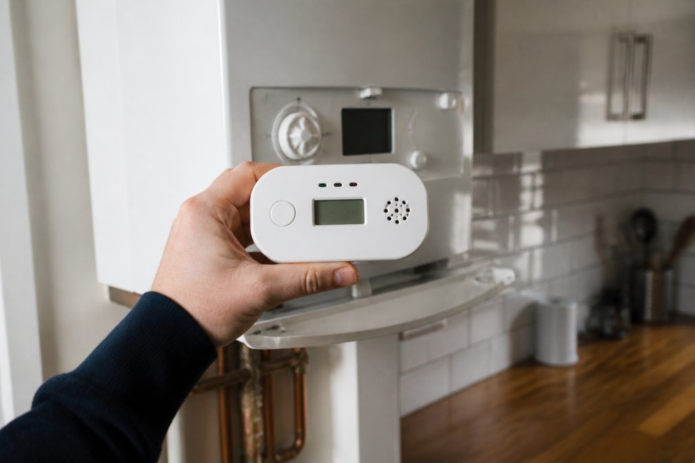 A close-up of a hand holding a portable carbon monoxide detector next to a gas boiler in a modern kitchen, showing the digital display reading zero