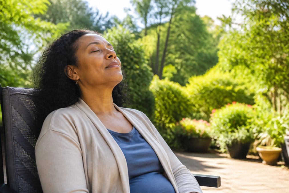 Caregiver sitting peacefully outdoors in natural sunlight with eyes closed, shoulders relaxed, surrounded by greenery, taking a genuine break