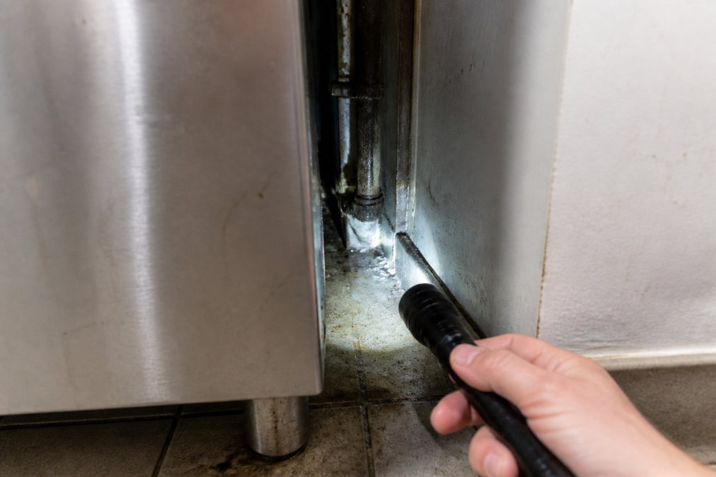 Close-up of a commercial kitchen corner showing a stainless steel unit pulled slightly away from the wall, revealing a dark gap with pipework and a small torch beam illuminating the space