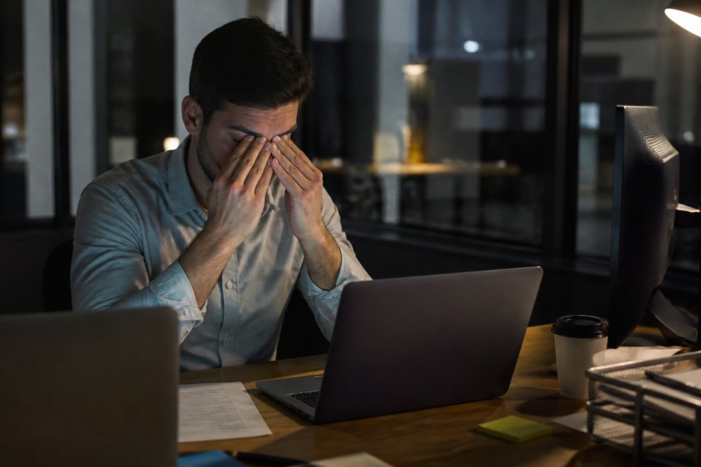 Professional showing signs of digital fatigue at desk with multiple screens, rubbing tired eyes with visible exhaustion, dim office lighting emphasizing fatigue